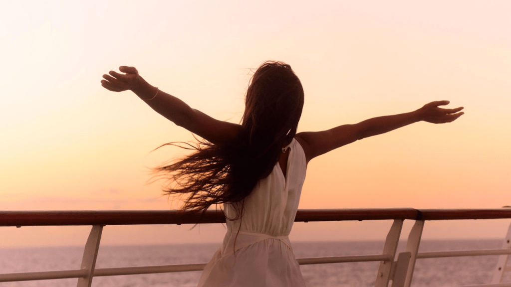 Woman with open arms in front of the railing of a cruise ship as the sun sets.